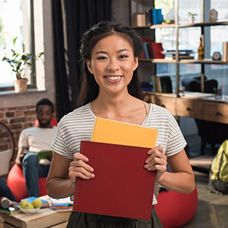 asian female student with folders smiling at the camera with a blurry background of a study hall